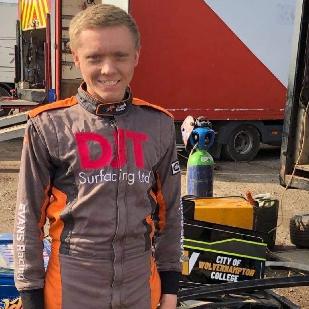 Engineering apprentice Lewis Evans, wearing a grey boiler suit in front of his stock car which displays the college's logo in gold and white writing on a black background