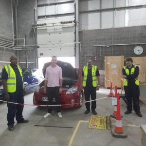 BBC's The One Show presenter Joe Cowley, wearing a pink shirt, and three students in high vis jackets in front of a car in the electric vehicle and green technologies centre at the Wellington Road campus