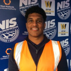 Photograph of former rail cecntre Richard Thomas-Robinson wearing a fluorescent orange jacket and standing in front of a sign showing the logos of the college and is employer, National Infrastructure Solutions