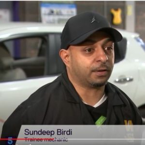 Electric vehicle student Sundeep Birdi wearing a black cap and black jacket with the college logo, talking to a reporter on 5 News