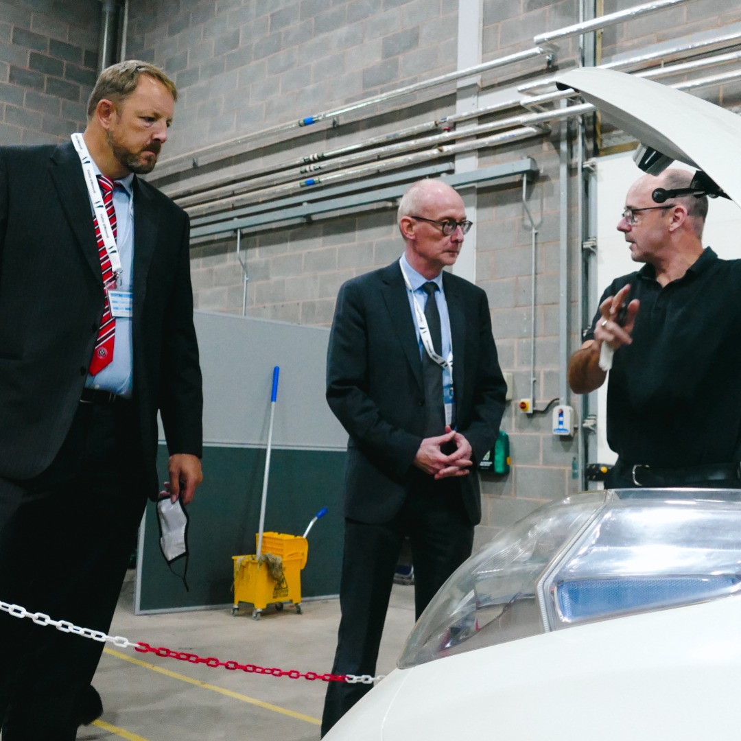 Toby Perkins, shadow minister for further education (left) wearing a dark suit, pale blue shirt and red striped tie, with Pat McFadden, MP, wearing a dark suit, plae blue shirt and dark tie, talking to a tutor in the motor vehicle workshop at the Wellington Road campus