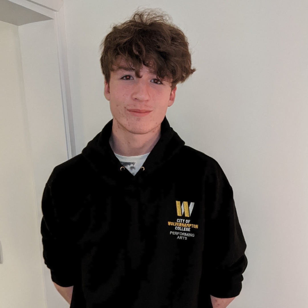 Performing arts student Josh Molloy, standign in front of a white background, wearing a black sweatshirt with the white and yellow college logo on the left side.