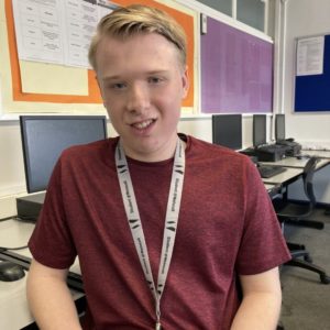 Business student Elliot Devlin, wearing a burgundy t-shirt and a grey colleg lanyard, sitting in front of a PC in a classroom