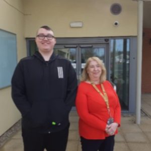 Student Mason Danbury on the left, wearing black trousers and a black hoodie, with his support worker standing on his left, wearing an orange top with a yellow college lanyard