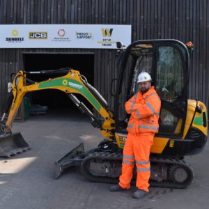Groundworks trainer Nathon Kerr, wearing an organe overall and a white hard hat standing in front of a digger at the college's construction centre