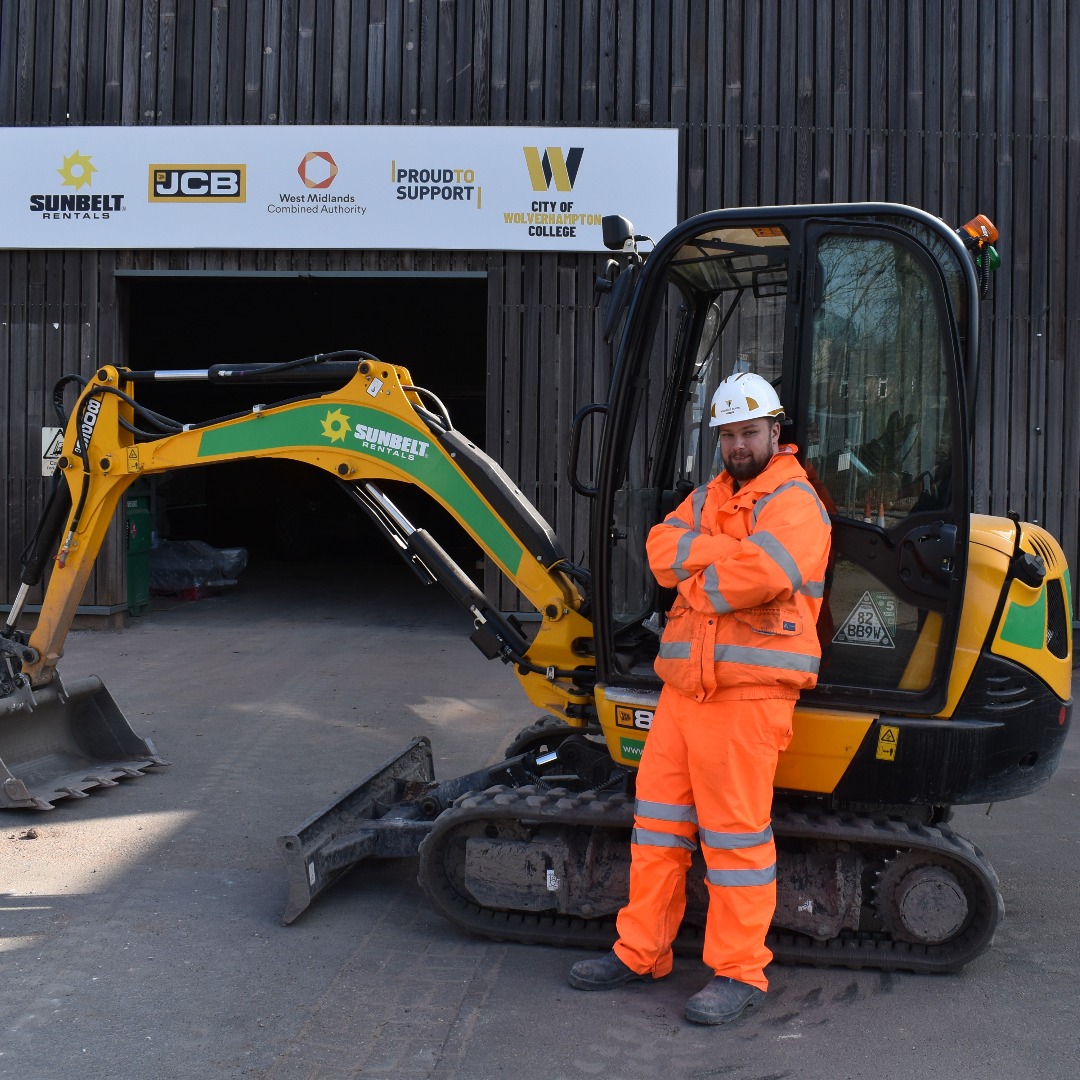 Groundworks trainer Nathon Kerr, wearing an organe overall and a white hard hat standing in front of a digger at the college's construction centre