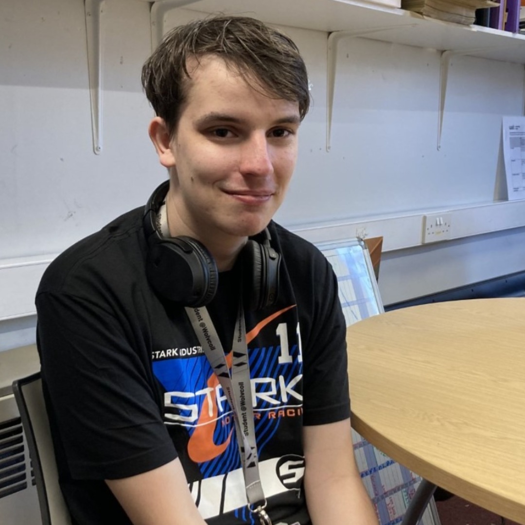 Music technology student Alex Pavey, wearing a black t-shirt with a multi-coloured pattern on the front - sitting by a table in a classroom