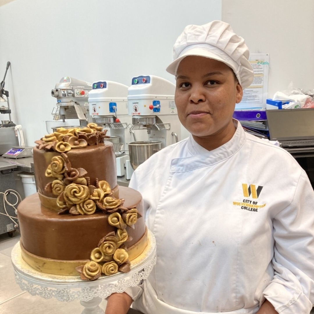 Bakery student Comfort Komape wearing a white overall and hat, pictured with a two-tier chocolate celebration cake, decorated with gold flowers, that she made