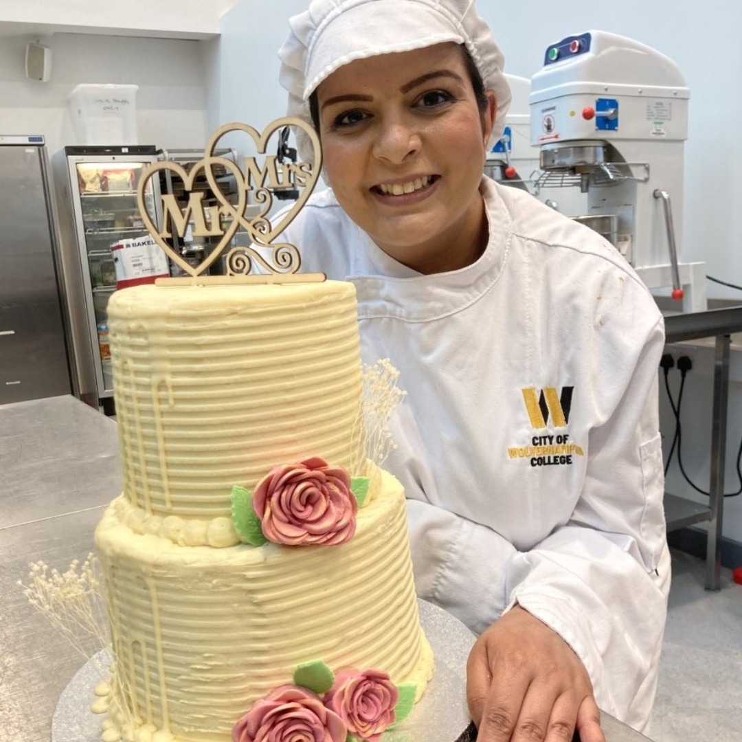 Bakery student Preeti Jhalli, wearing a white overall and white hat, with a two-tier wedding cake that she had made and covered with white chocolate and handmade flowers