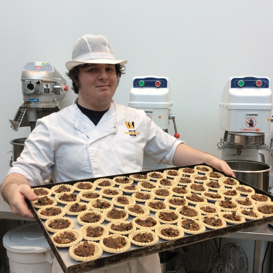 Bakery student Jack Stevens wearing a white overall and white hat, holding a large tray of mince pies in the bakery kitchen at the Wellington Road campus