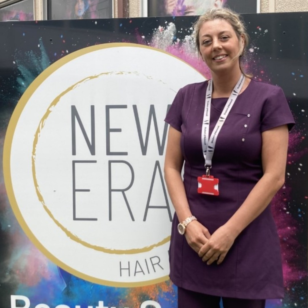 Beauty student April WOlverson, wearing a purple uniform, standing by the sign for the New Era hair & beauty salons. The logo is a white circle with a yellow edging with the words New Era in the centre in grey capital letters