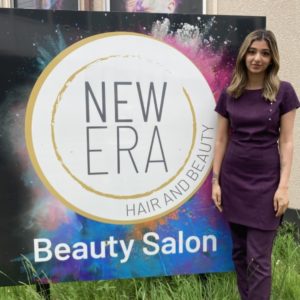 Beauty student Ayesha Paul, wearing a purple uniform, standing in front of a sign advertising the new Era beauty salon