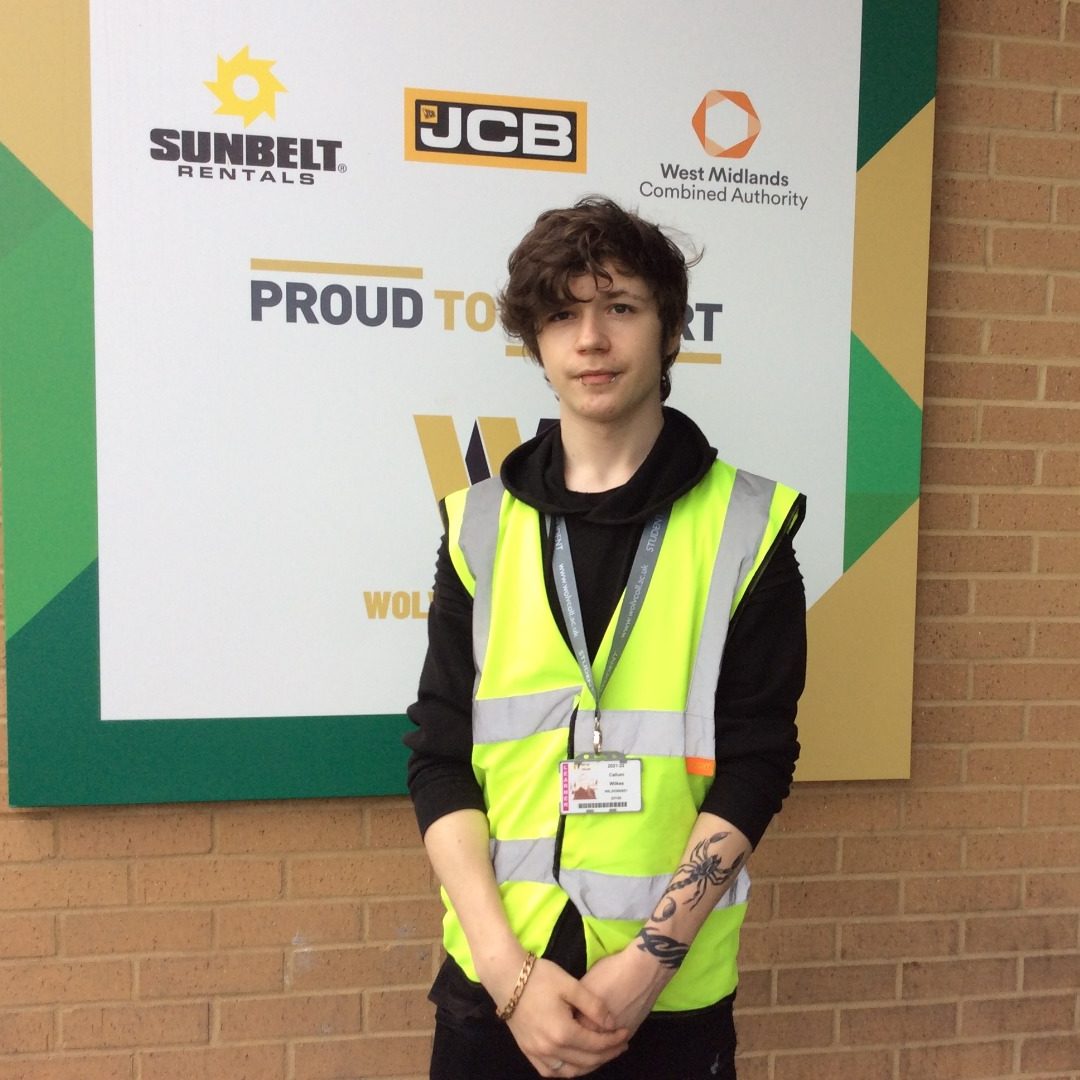 Student Callum Wilkes, wearing a black top and a yellow florescent jacket standing in front of a sign outside the college's construction centre