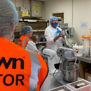 Cheft from Dawn Foods in Evesham, wearing a white overall land blue hair net, talking to students in the kitchen during a visit to the factory