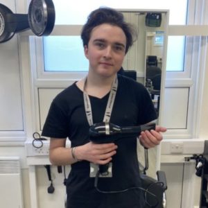 Barbering student Liam Davis wearing a black top and holding a hairdryer, pictured in the college's training salon