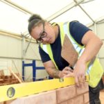 Female bricklaying student wearing a black t-shirt anf hi-vis jecket laying a spirit level on the top of a brick wall