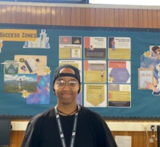 Student Dalila Madiera wearing a black cap and black top, standing in front of a notice board