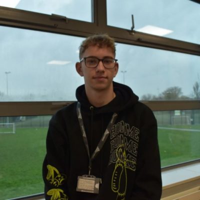 Sport student Lewis Smith wearing a black hoodie and a college lanyard, standing in front of a window overlooking the sports pitch