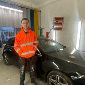 Former automotive student Tom Coates wearing an orange hi-vis jacket standing in front of a balck car in the workshop ar dealership Greenhous where he's now working