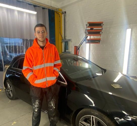 Former automotive student Tom Coates wearing an orange hi-vis jacket standing in front of a balck car in the workshop ar dealership Greenhous where he's now working