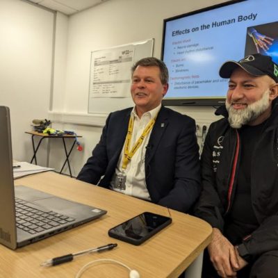 Andy Moore (left) wearing a dark jacket, white shirt and yellow college lanyard, with badar Zaman (right) wearing a dark jacket and cap, sitting in front of a laptop recording the podcast