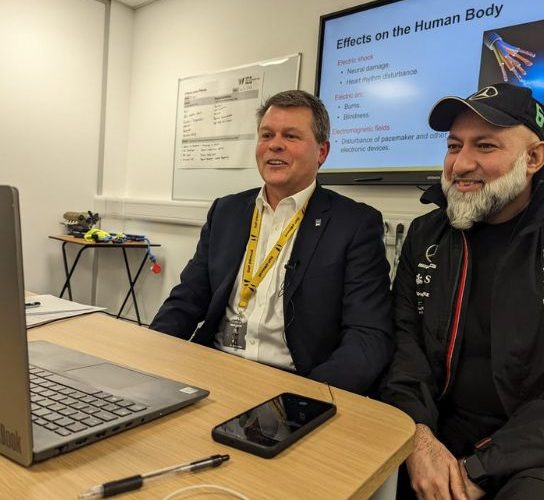 Andy Moore (left) wearing a dark jacket, white shirt and yellow college lanyard, with badar Zaman (right) wearing a dark jacket and cap, sitting in front of a laptop recording the podcast