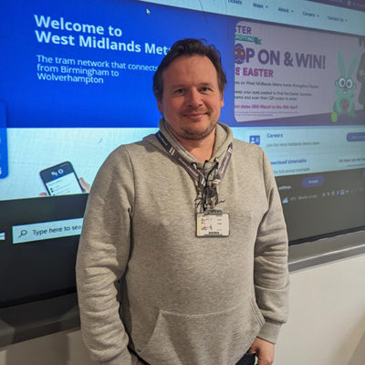 Colin Todd, wearing a grey top, standing in front of a display board with information about Midlands Metro careers