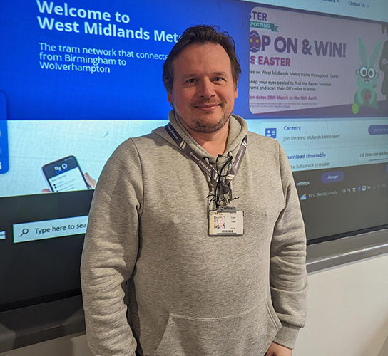 Colin Todd, wearing a grey top, standing in front of a display board with information about Midlands Metro careers