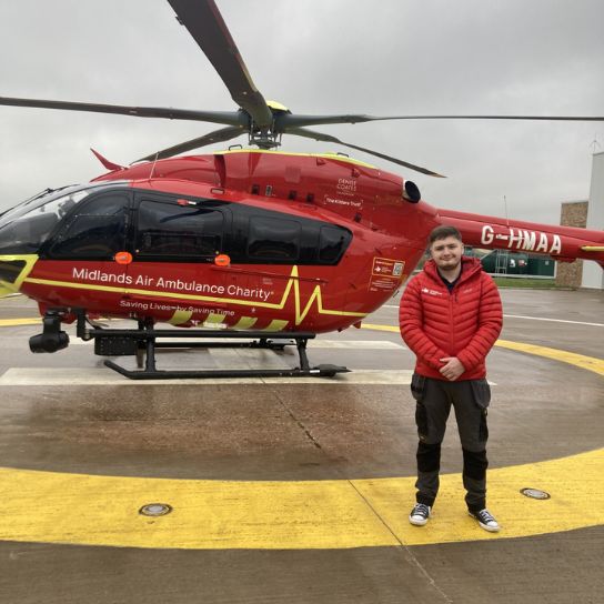 Apprentice wearing a red jacket standing in front of a red Midlands Air Ambulance Charity helicopter