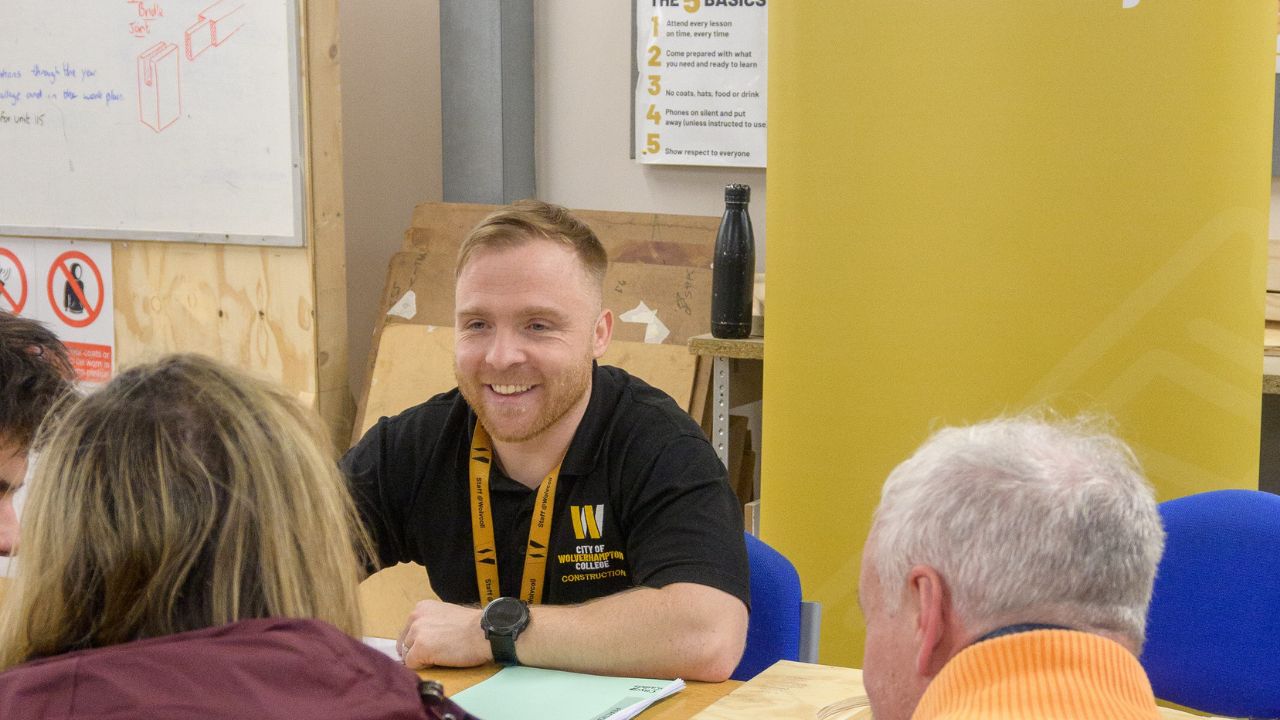 A tutor wearing a college-branded top and lanyard talking to students at an open evening